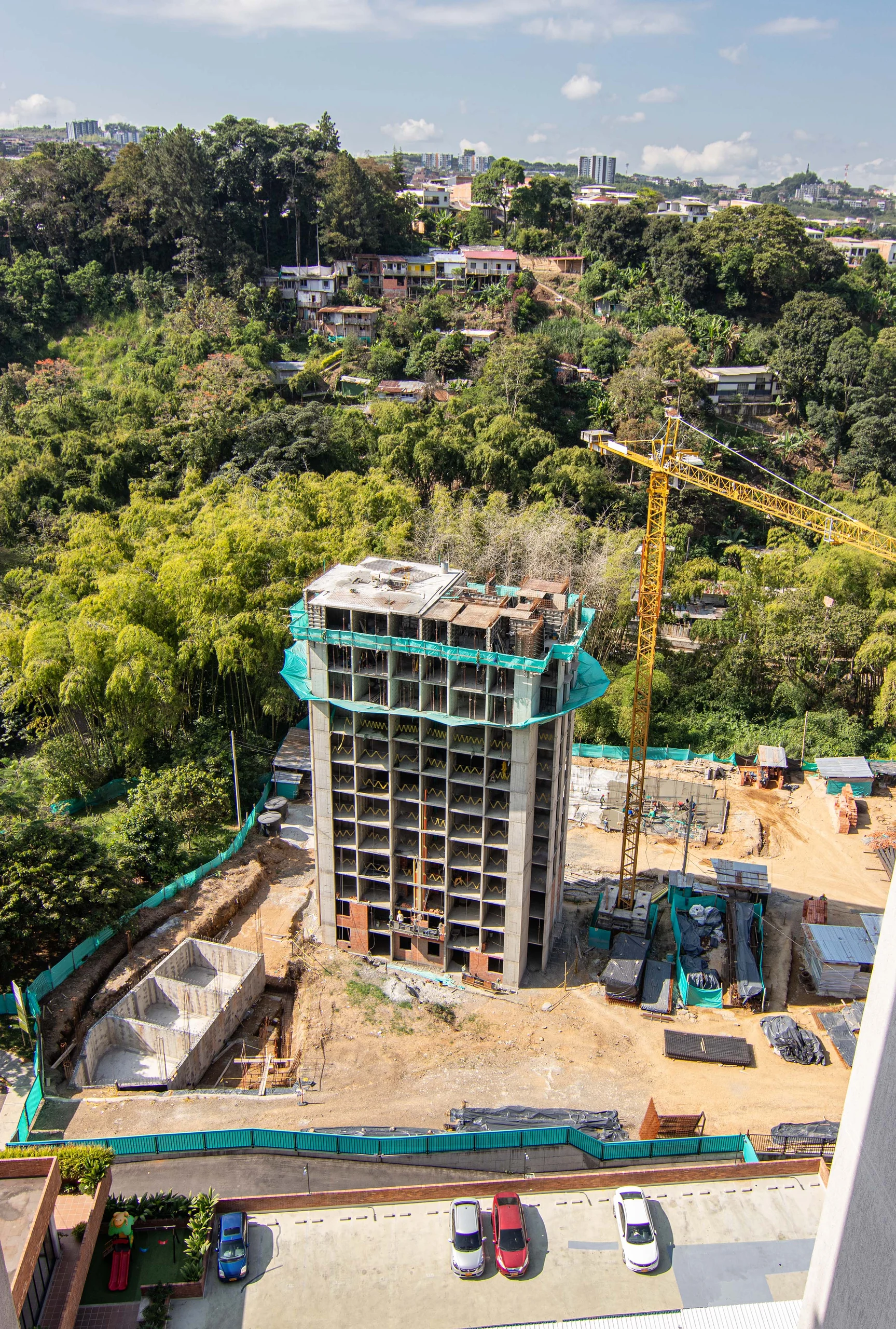 The tower under construction rises over the guadua hillside of Pereira; the crane and the stepped platforms reveal how the project settles onto the slope.
